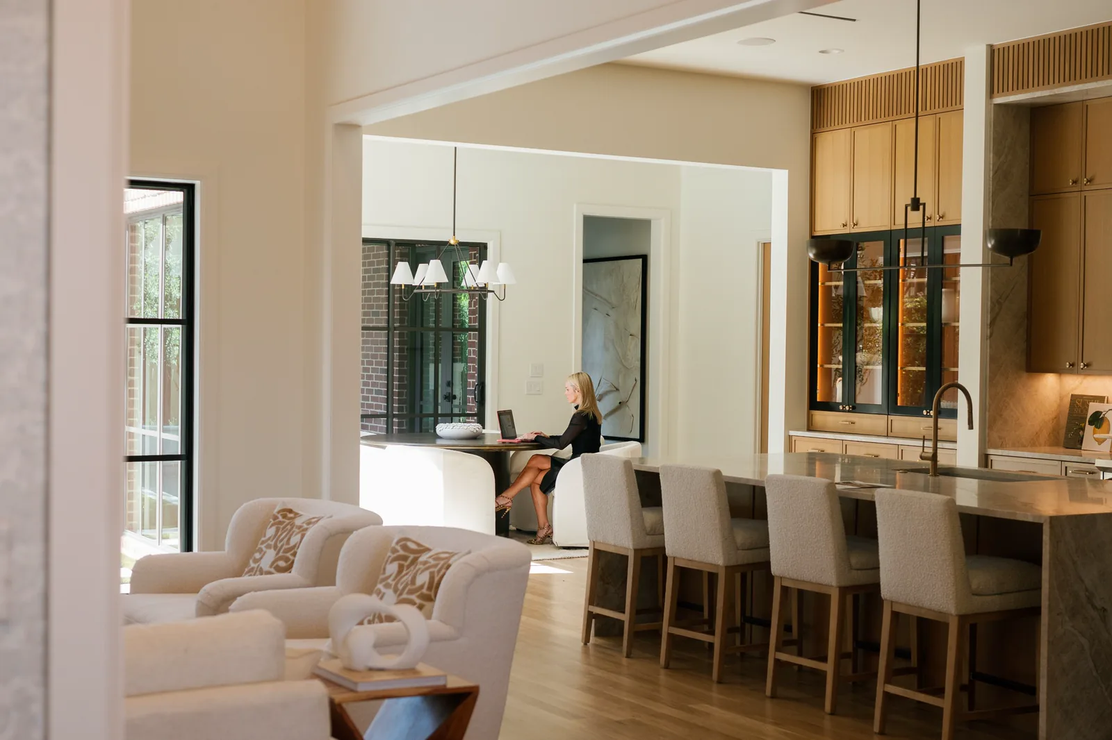 Open great room with white armchairs, a kitchen island with bar stools, and a dark-framed wet bar beyond.