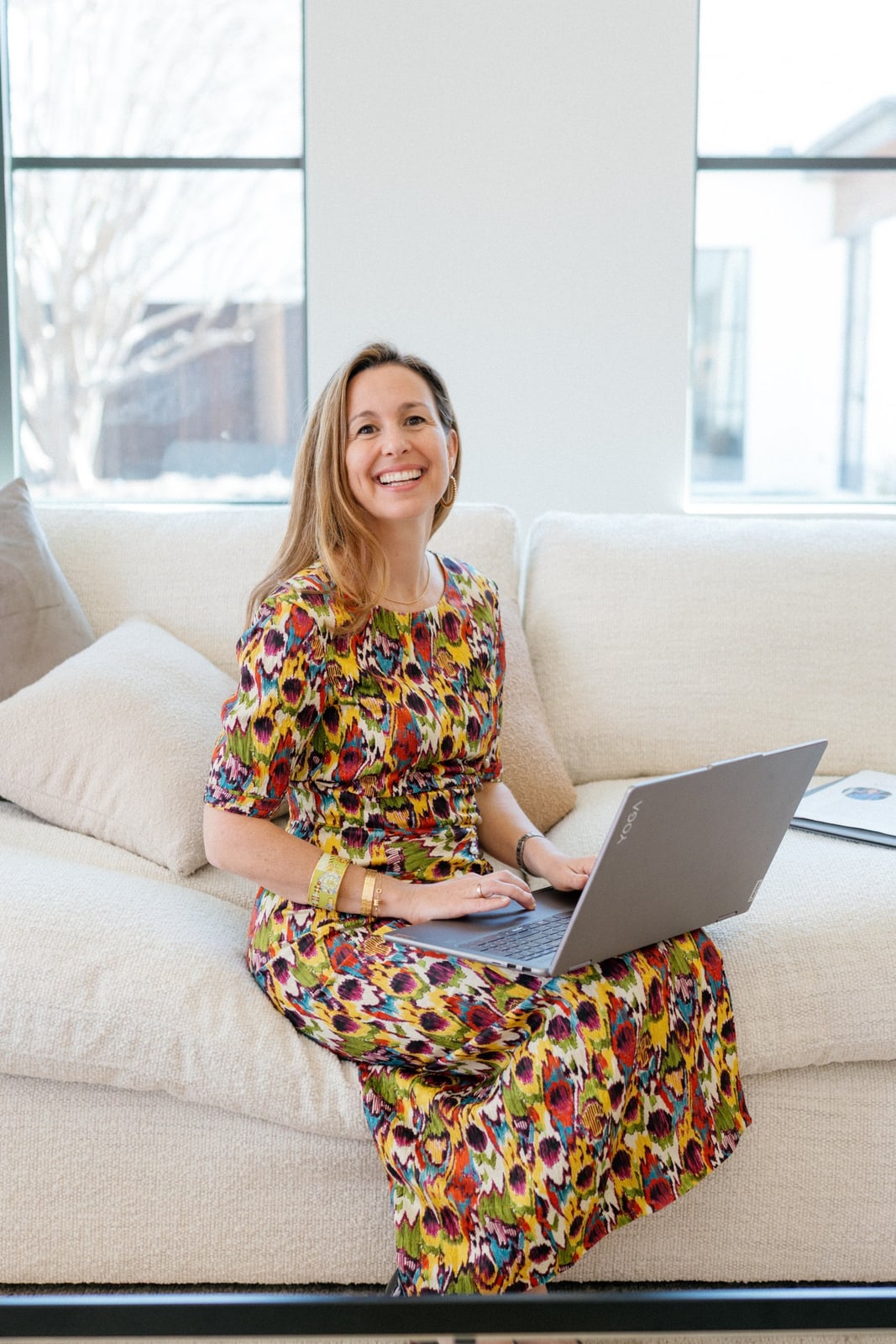 Smiling woman in a floral dress working on a laptop on a white sofa.
