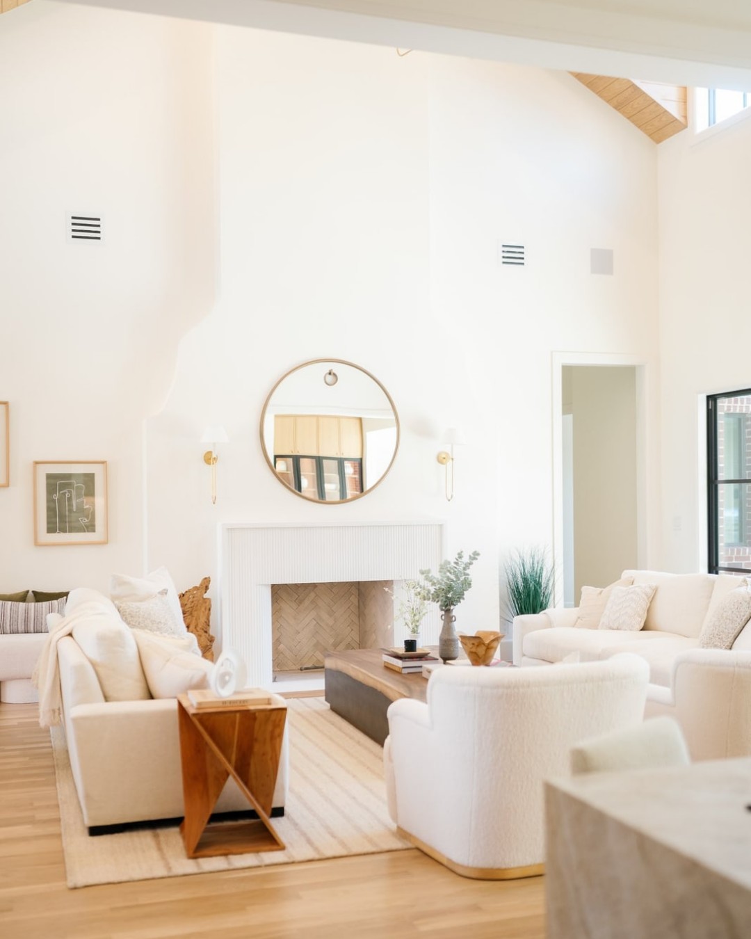 Two-story white living room with a fireplace, round mirror and cream curved sofas.