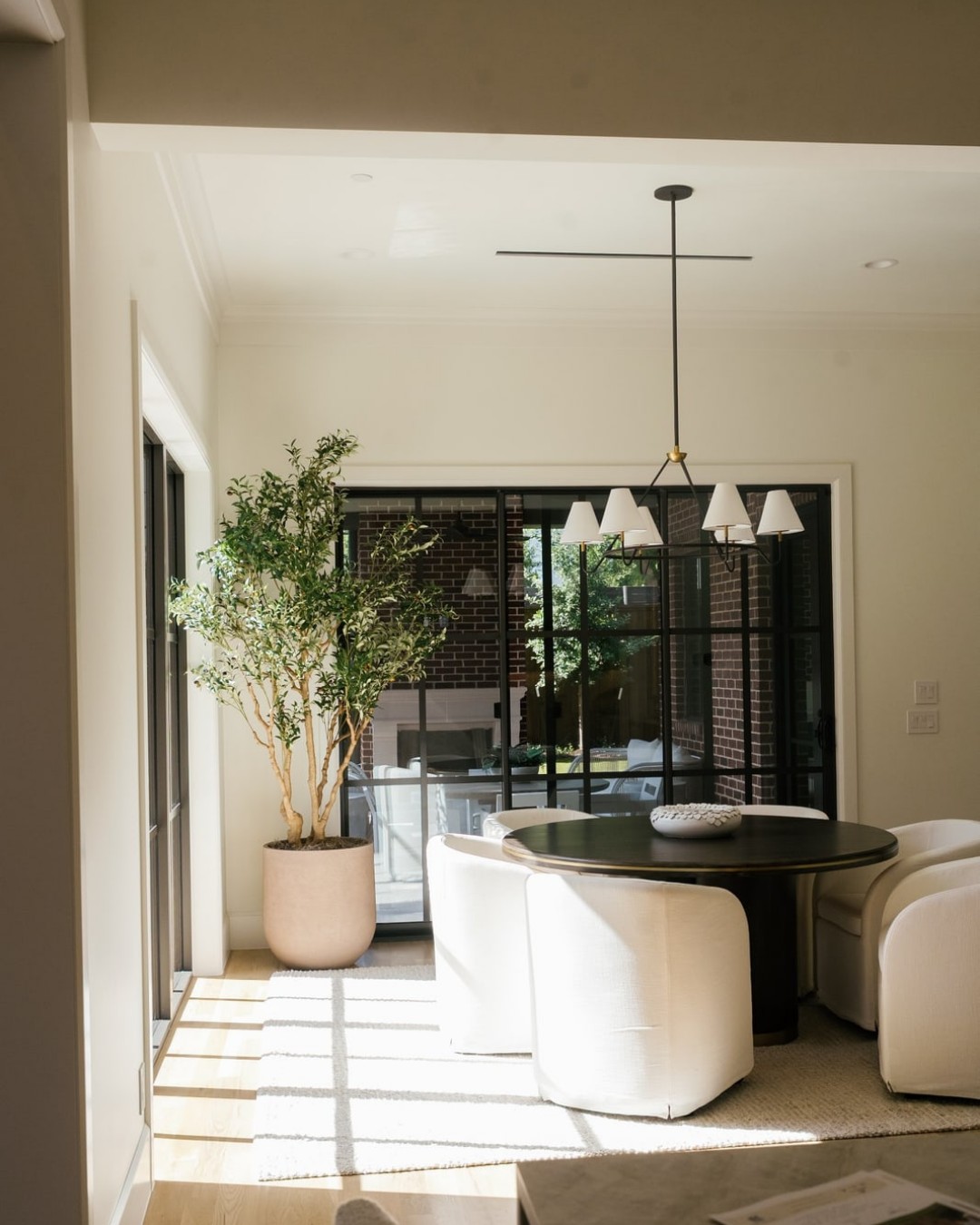 Sunlit breakfast nook with a round table, white chairs, a pendant light and a potted tree.
