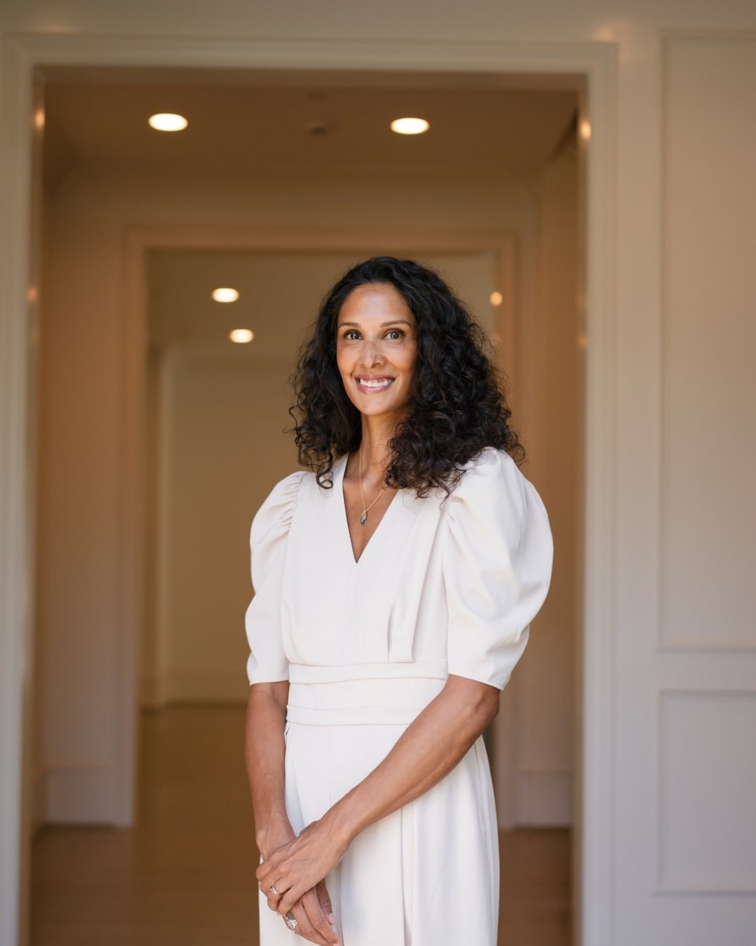 Woman in a white dress standing in a long hallway with recessed lighting.