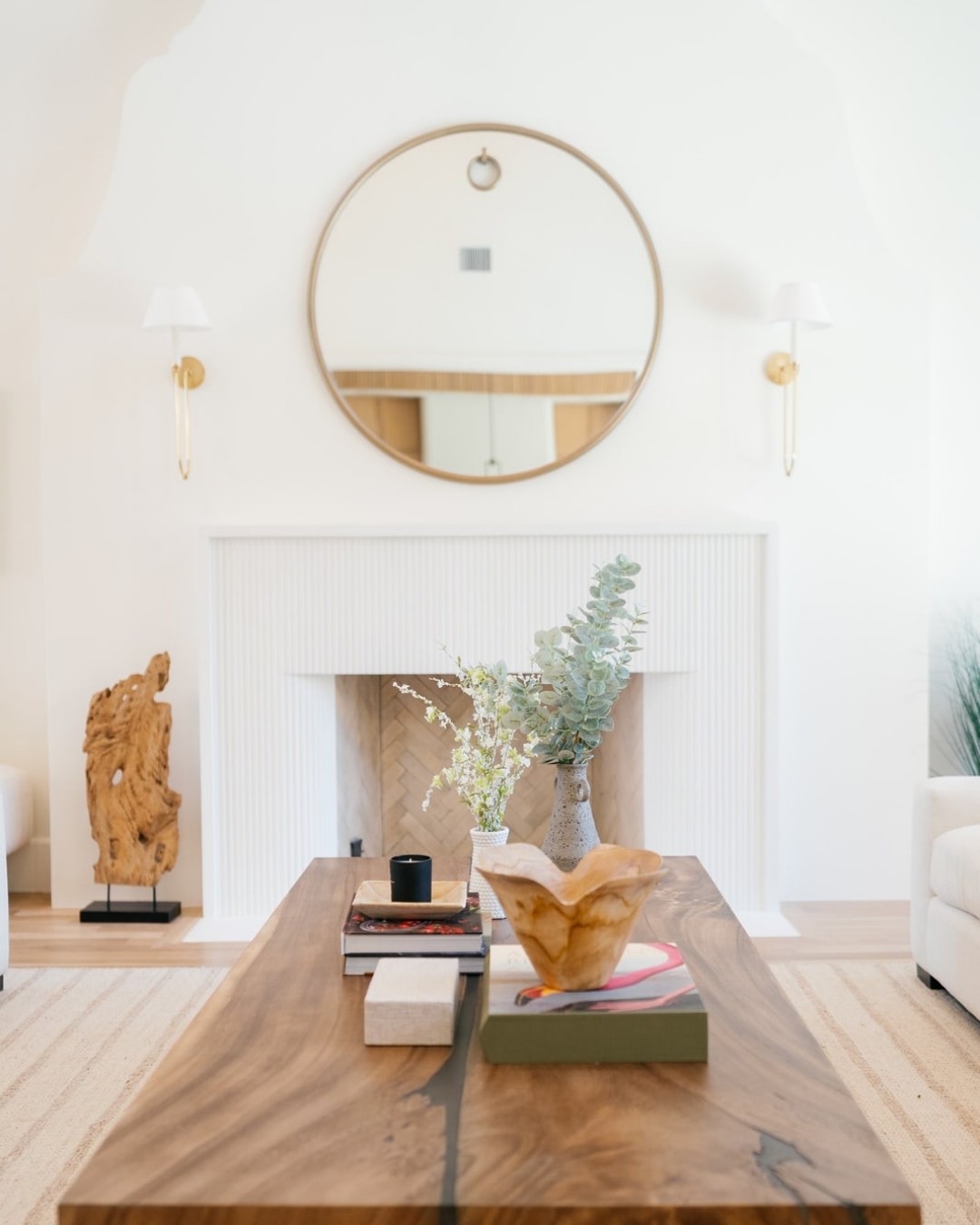 White fireplace under a round brass mirror with a live-edge wood coffee table.
